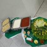 Duas mesas circulares, uma ao lado da outra, contendo saladas, molhos e massas em buffet infantil